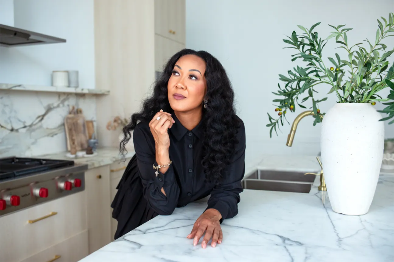 Woman with long curly hair wearing black blouse leaning on a marble kitchen countertop.